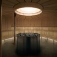 A modern circular sauna with wooden benches lining the walls, a central metal stove filled with rocks, and a gong hanging on the right. Natural light enters from a round ceiling window.
