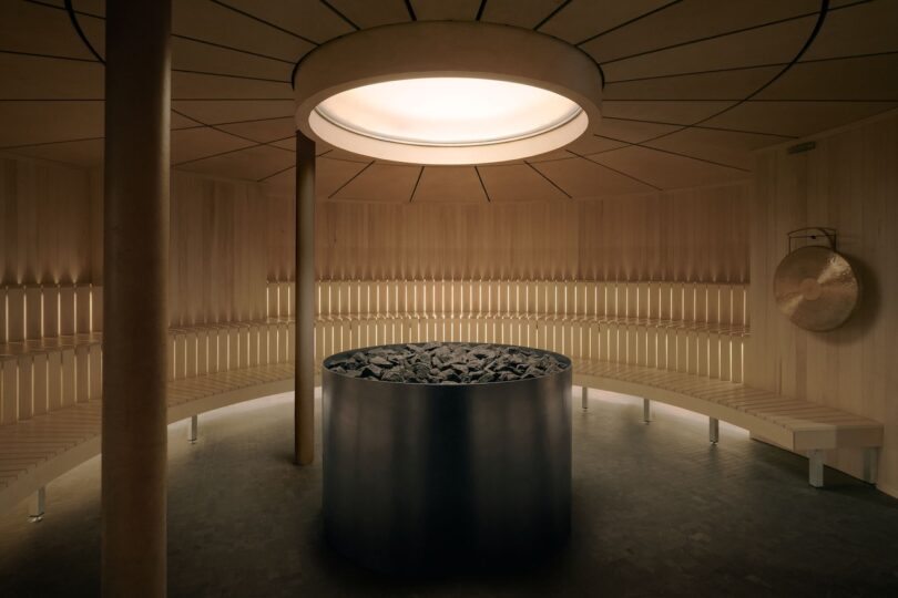 A modern circular sauna with wooden benches lining the walls, a central metal stove filled with rocks, and a gong hanging on the right. Natural light enters from a round ceiling window.