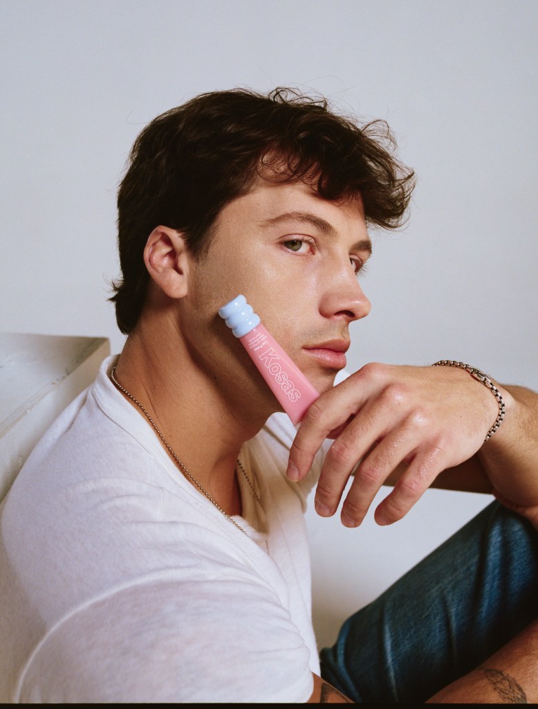 A man with green eyes and brown hair holds a pink Kosas Lip Oil tube to his cheek.