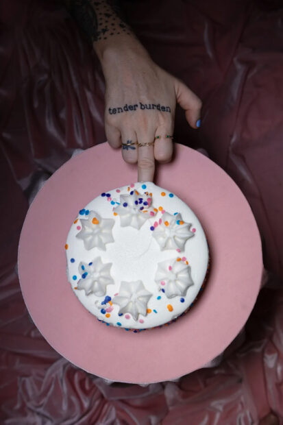 A photograph by Emily Peacock of a tattooed hand sticking its middle finger into a white cake.