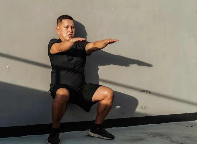 Middle-aged Southeast Asian man holds a wall sit with arms raised, showing control, endurance and core activation during bodyweight isometric training.