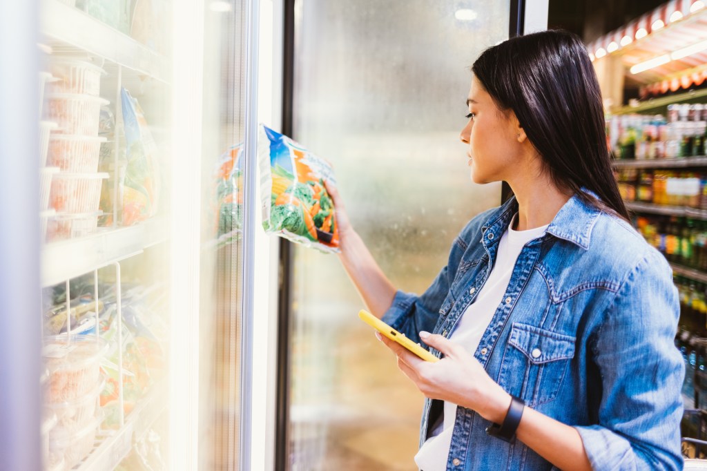 A young woman holding a phone in one hand and a bag of frozen vegetables in the other.