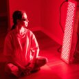 A photo of a woman sitting on the floor next to a red light therapy device in her home.