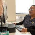 A nurse sitting at a computer smiles while talking to a patient at the Student Health and Wellness Center