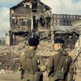 Two soldiers in green uniforms with their backs to camera, a bomb building is visible in front of them with debris all across the road