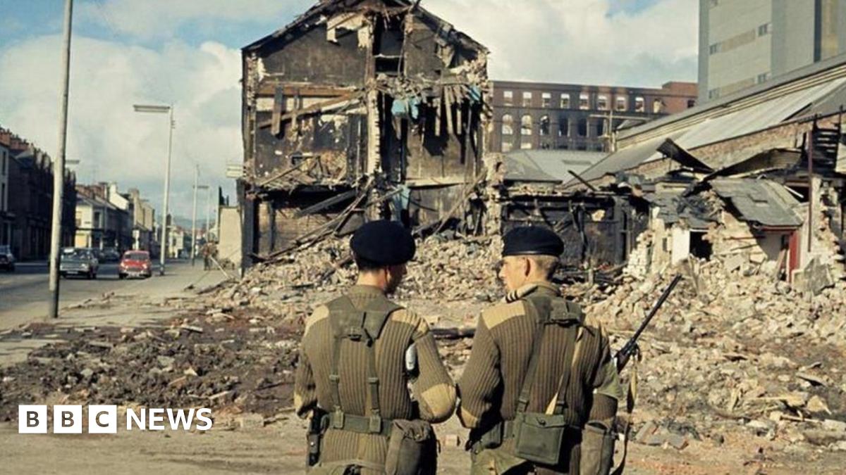 Two soldiers in green uniforms with their backs to camera, a bomb building is visible in front of them with debris all across the road
