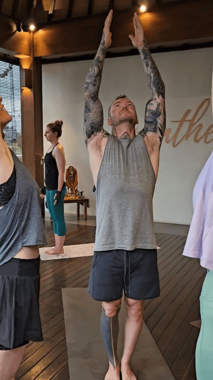 Group of people practicing yoga indoors, man with tattoos raising arms overhead in tree pose.