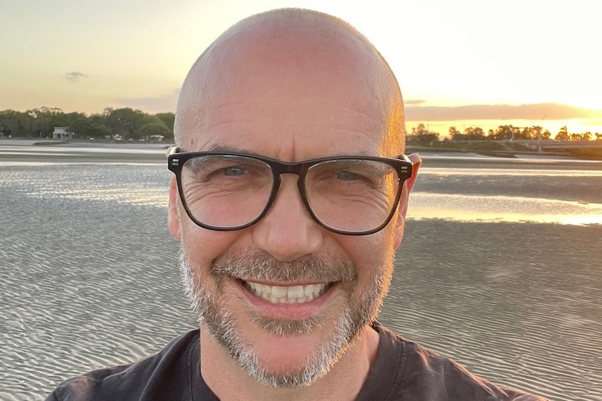 Man with glasses smiling at camera, while standing on a beach 