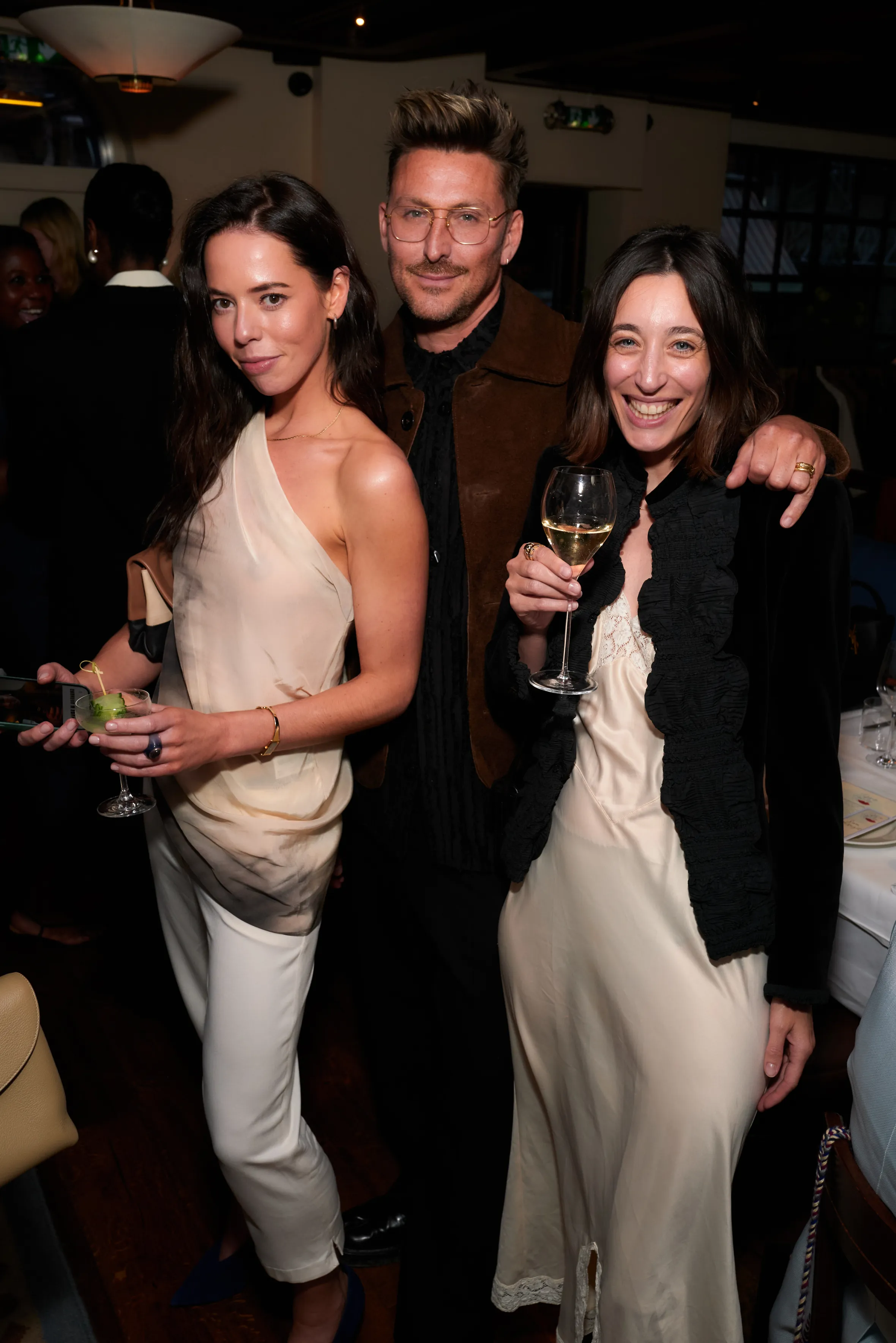 Three people standing and smiling, two women on either side of a man, holding drinks.
