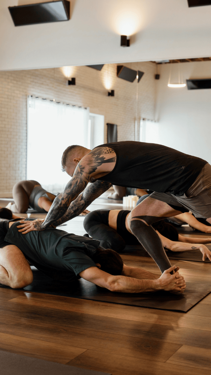 Yoga instructor assisting a student with a stretch in a bright, modern studio with wooden floors.