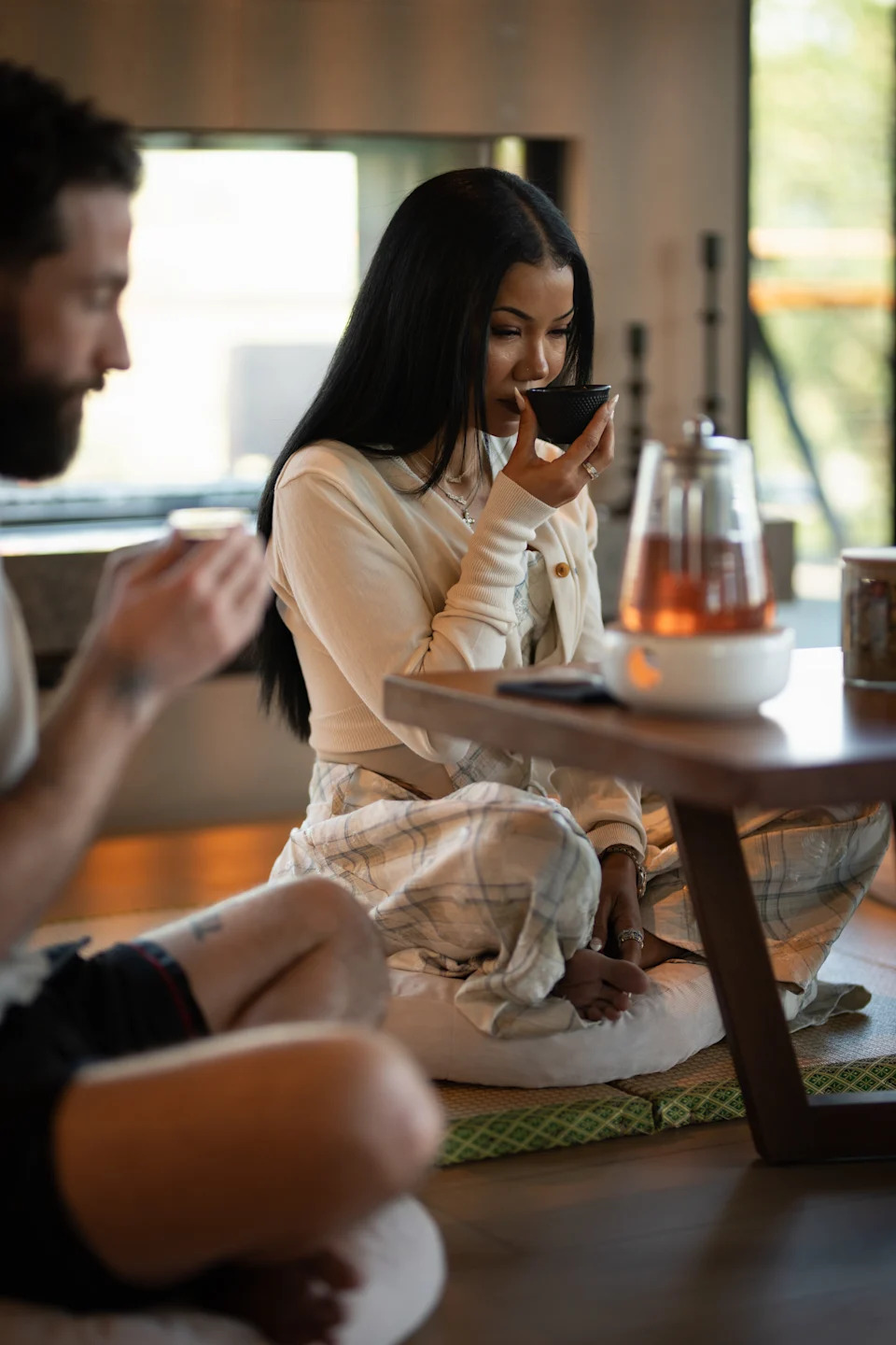 A woman with long black hair sits on a cushion, sipping tea, with a man sitting nearby. A teapot is on the table.