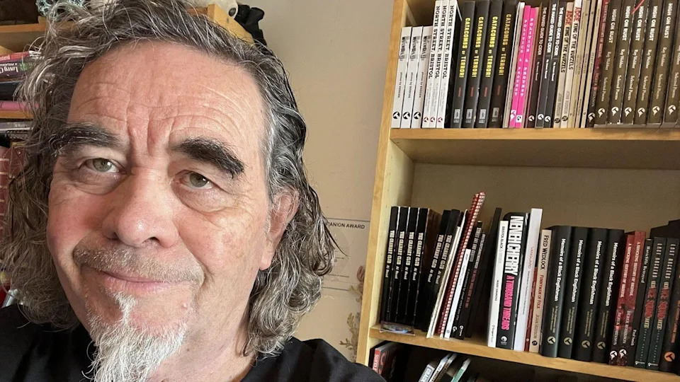 Richard has short curly hair and a beard and is looking at the camera on the left smiling. There is a bookshelf behind him of books.