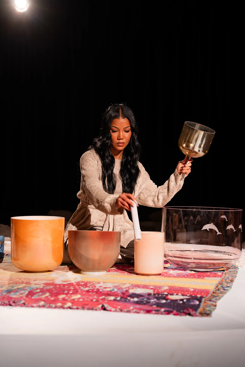 A woman with long black hair plays crystal singing bowls on a colorful rug in a dimly lit room.