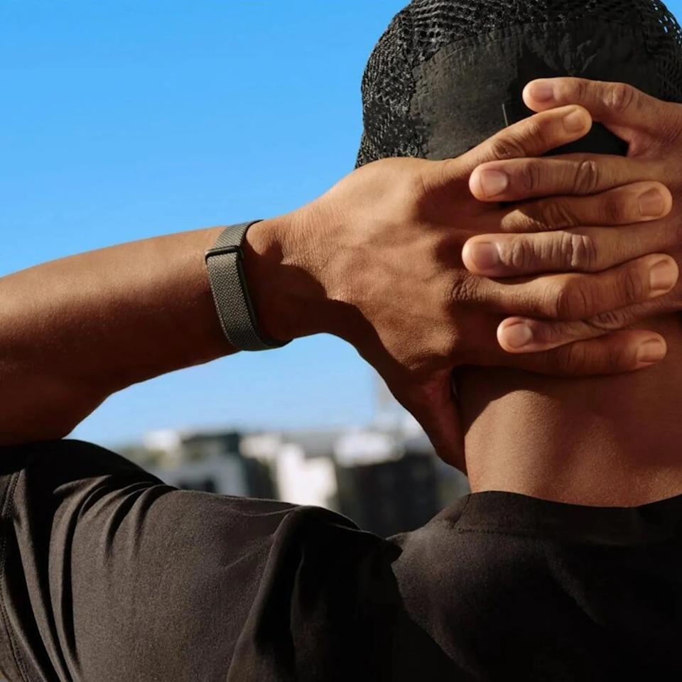 Person wearing a black mesh cap and black shirt with hands clasped behind their head against a clear blue sky.