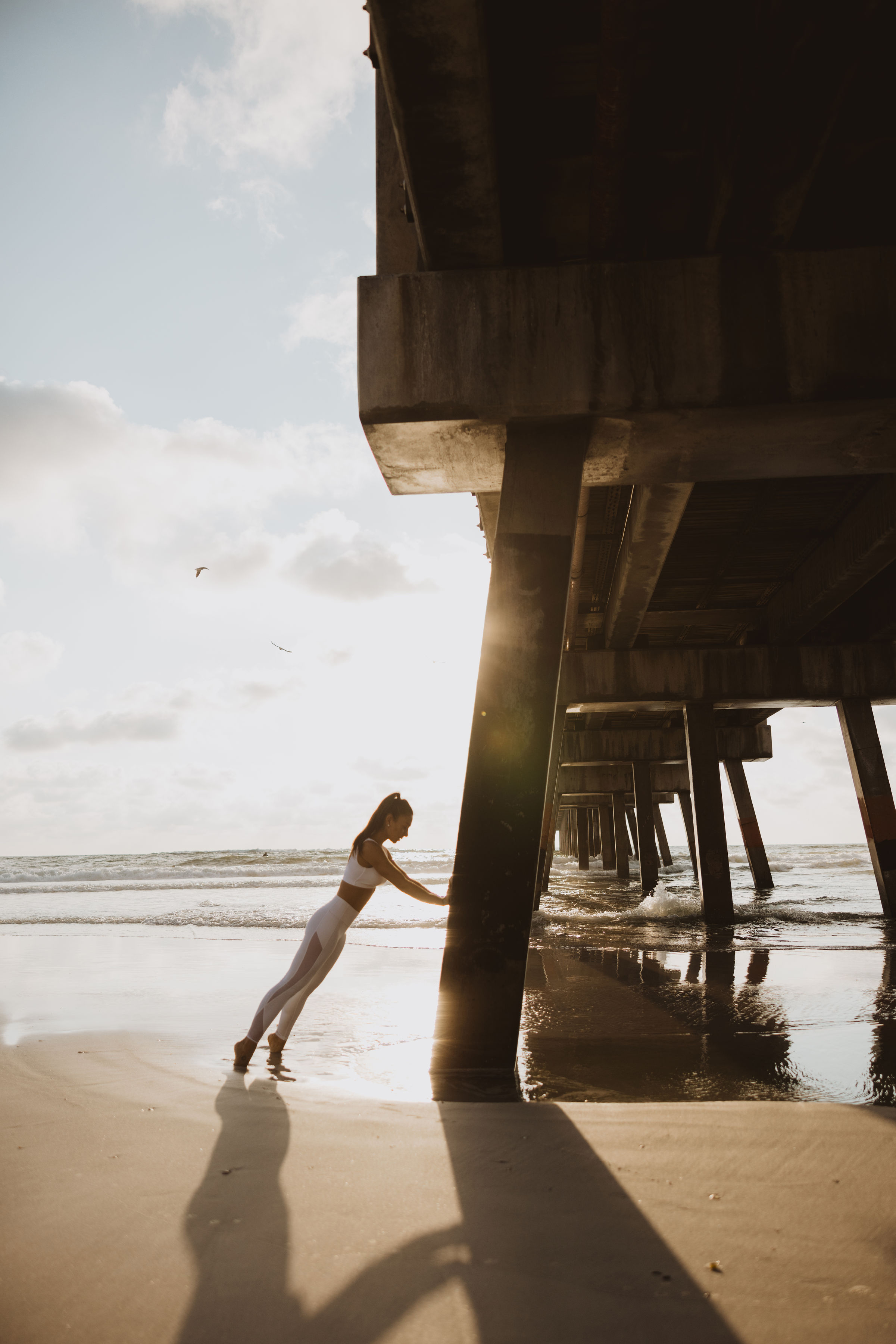 Person stretching legs against pier support on beach at sunrise or sunset.