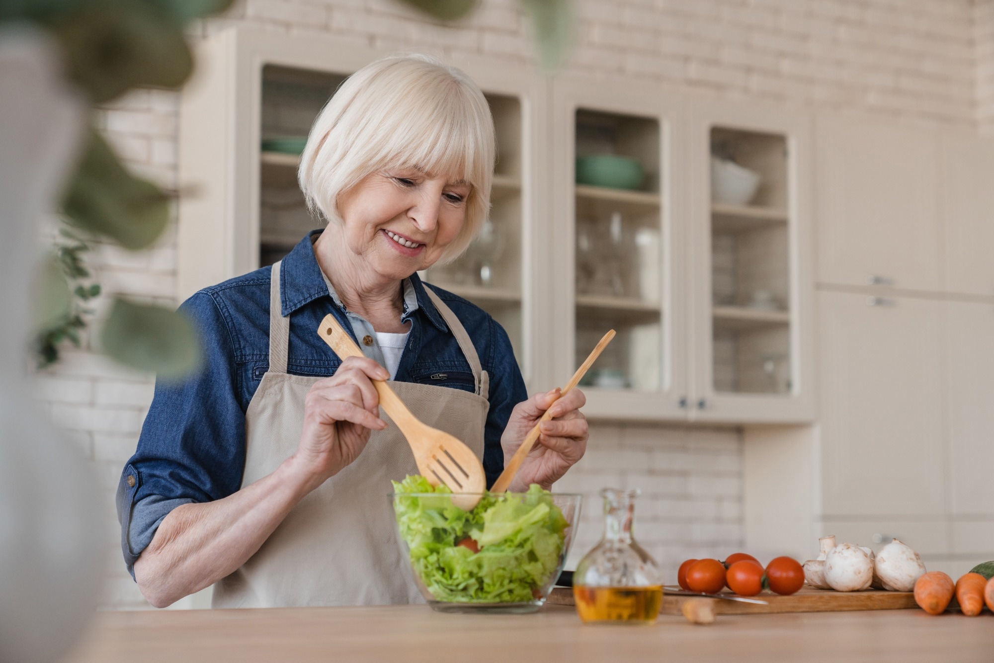 Older caucasian woman mixing ingredients in vegetable salad, cooking healthy food at home, preparing meal at home.