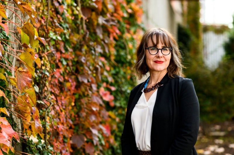 Professor Liz Bucar poses for a portrait in front of a wall of fall-colored foliage.