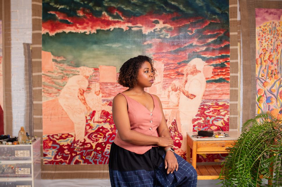 An artist sits in a studio in front of several large textile paintings featuring layered figurative scenes and vivid atmospheric colors.