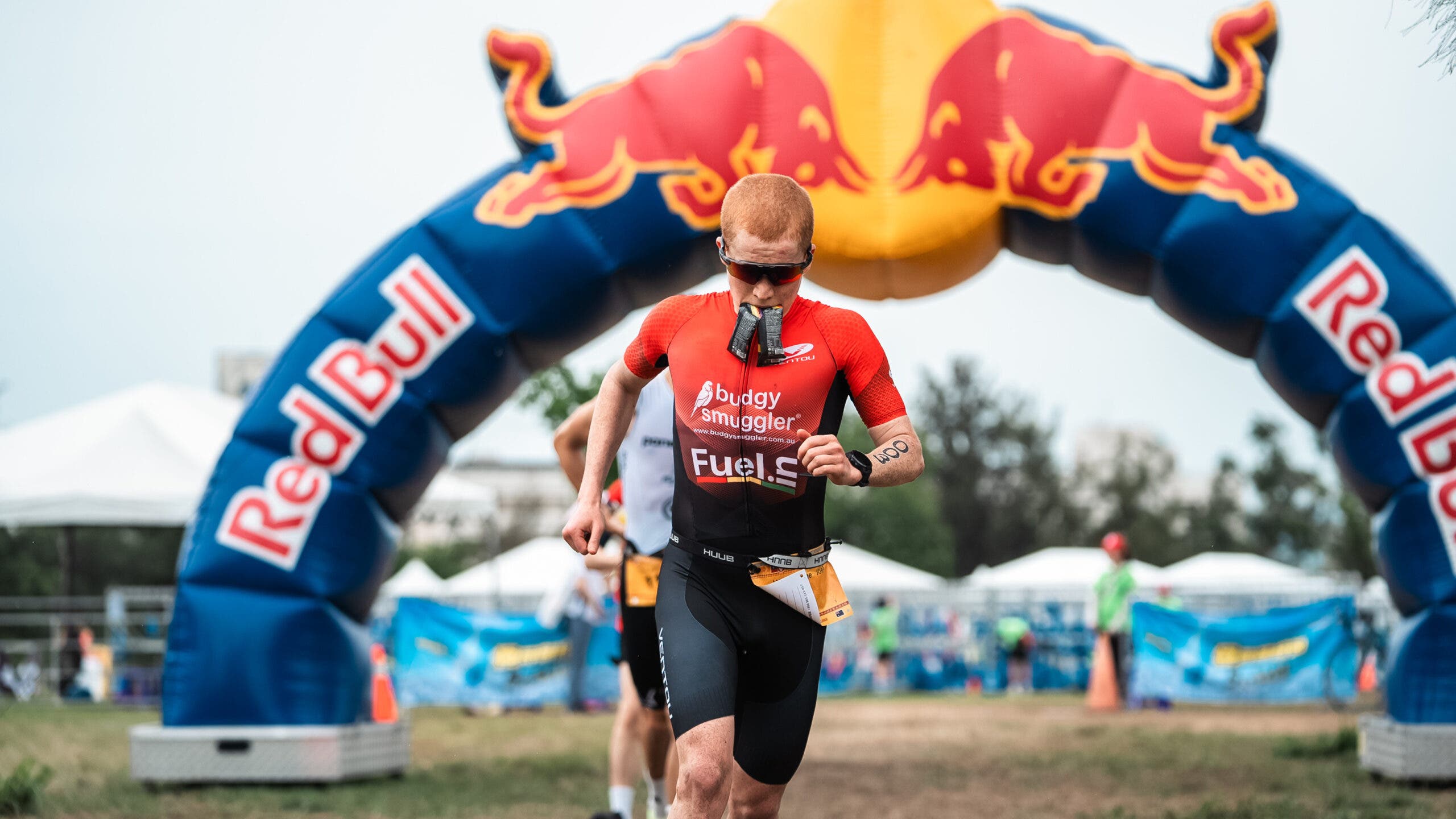 A triathlete runs under a Red Bull inflatable arch during a race with two gel packets hanging out of his mouth.