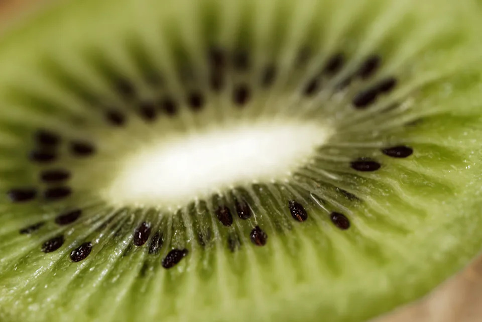 An extreme close-up of a kiwi fruit's internal structure, focusing on the dark brown seeds embedded in the succulent green and yellow pulp. The shot highlights the natural moisture, radiating fibers, and glistening texture of the fruit, making it perfect for organic food, wellness, and abstract nature themes.