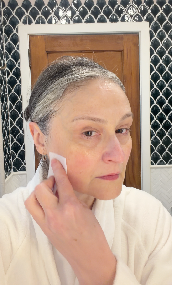 Person in a bathrobe applying skincare in a bathroom setting.