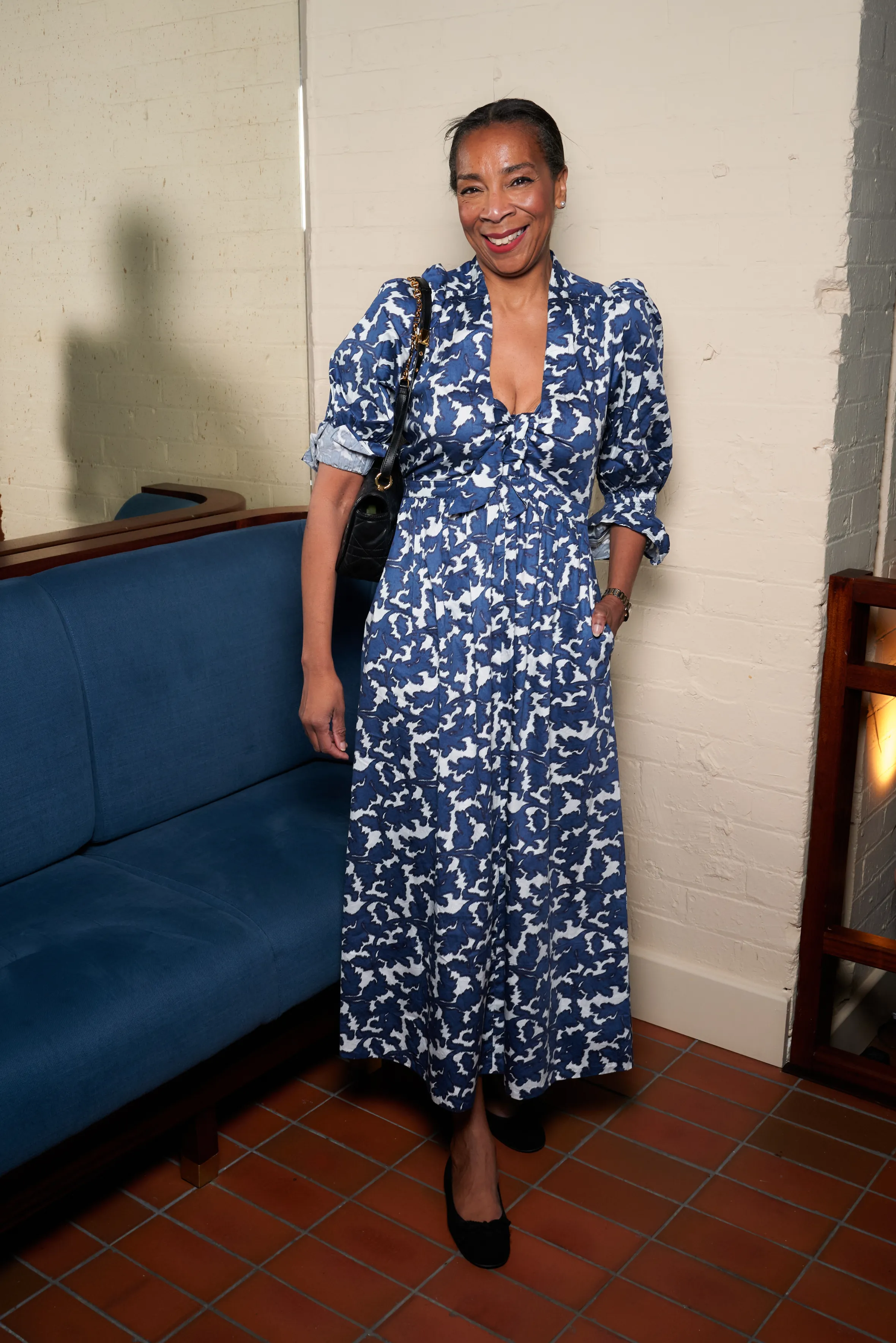 A woman in a blue and white patterned dress, with her hands in her pockets, smiling while standing next to a blue sofa.