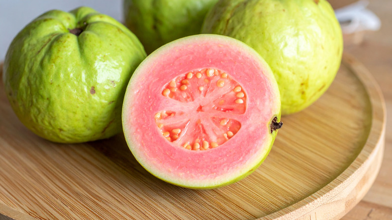 Guava sliced in half revealing pink flesh with small seeds the center and whole guavas in the background