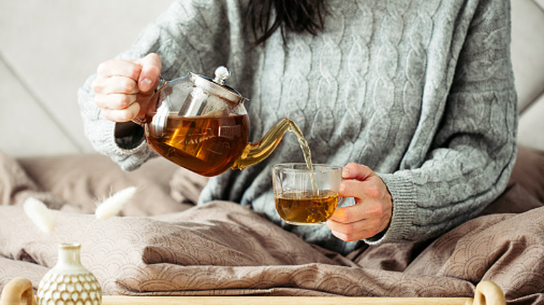 A woman pouring hot tea from a glass teapot.