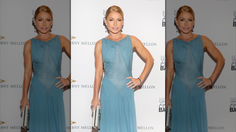 Kelly Ripa wears a blue dress and poses with a hand on her hip.