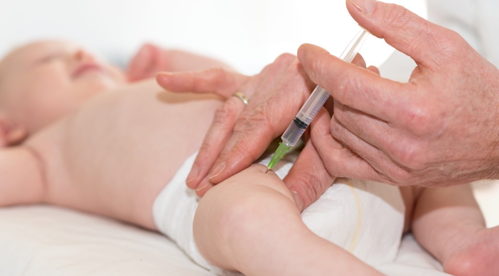 A Caucasian baby in a diaper receives a vaccine shot in the leg from an unseen healthcare professional.