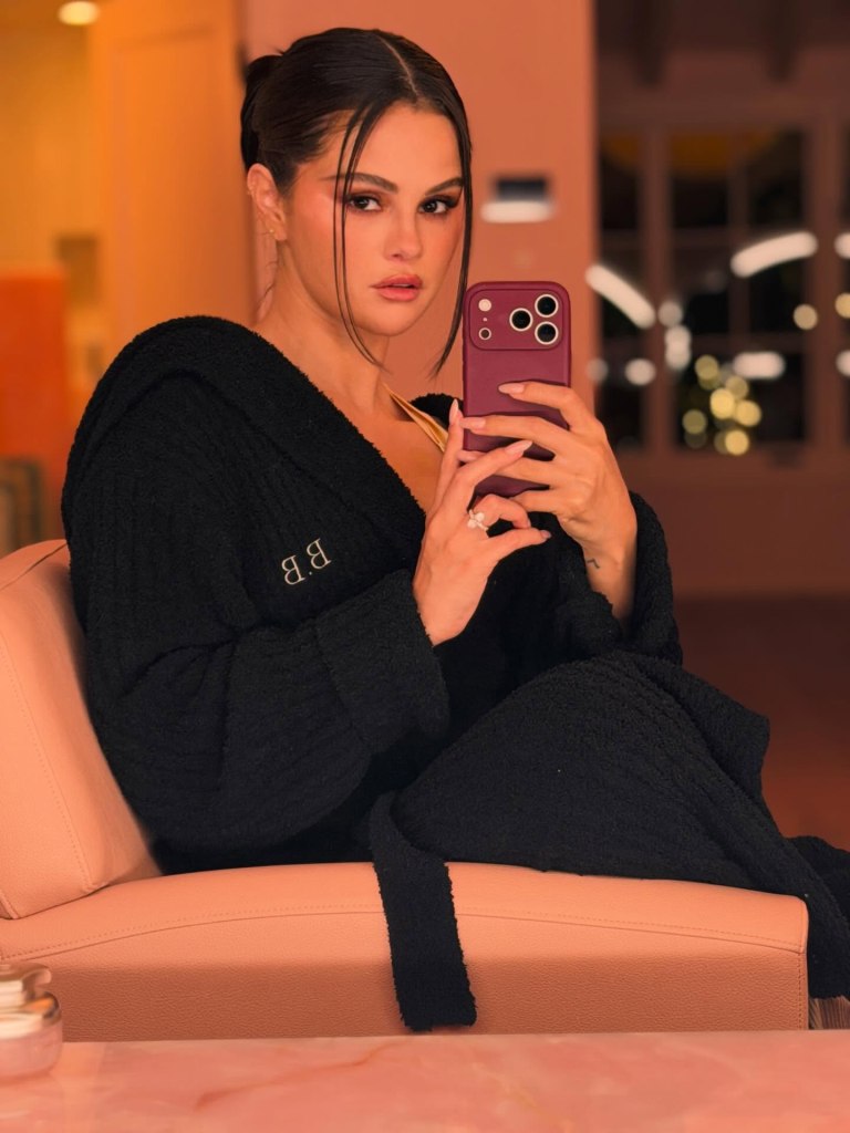 Selena Gomez in a black robe taking a selfie with a purple phone.