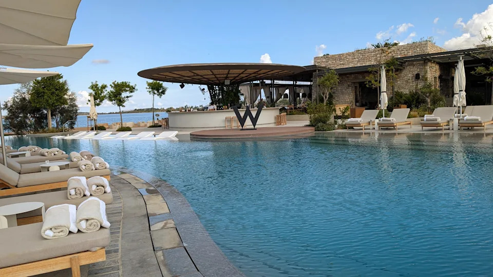 A wide landscape view of the large, modern outdoor infinity pool lined with lounge chairs and umbrellas at the W Costa Navarino resort in Greece.