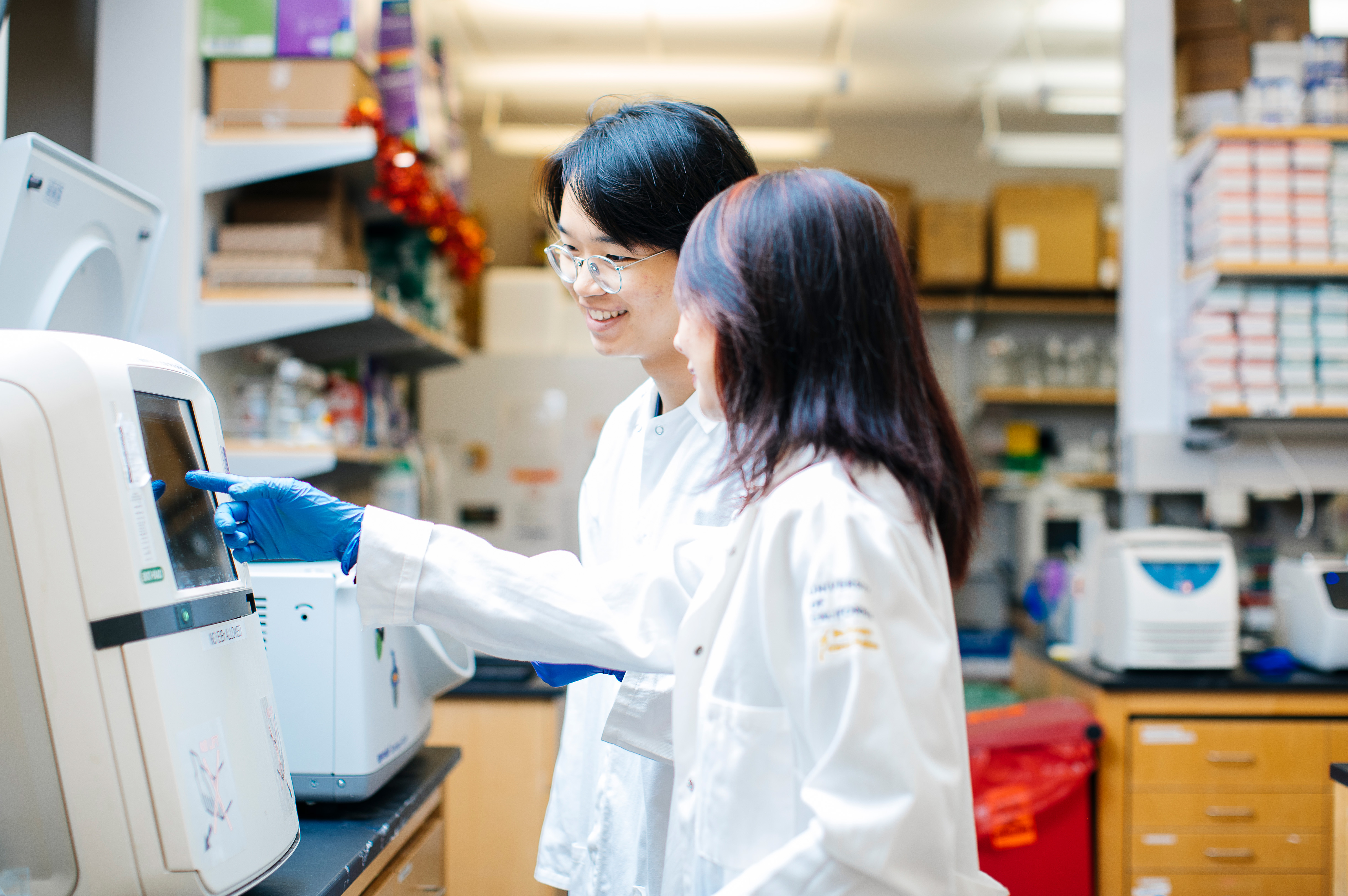 Elliot Kang with Lili Yang in the lab. Eliot Kang, with glasses, and Lili Yang wear lab coats and gloves as they review data on equipment in a scientific research lab.