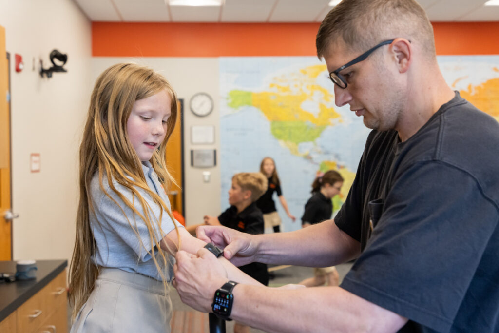 A man helps a young girl put on an activity tracker in a classroom with other children and a world map in the background.