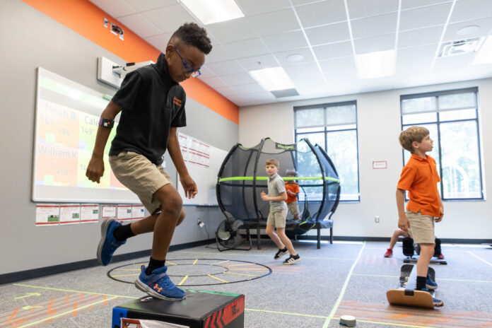 Three boys participate in physical activities in a classroom with exercise equipment and a trampoline in the background.