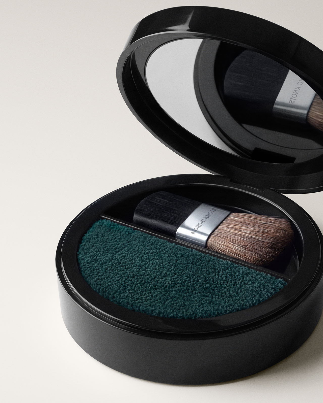 A black round compact case with a mirror, containing a green textured powder and a small flat makeup brush with brown bristles.