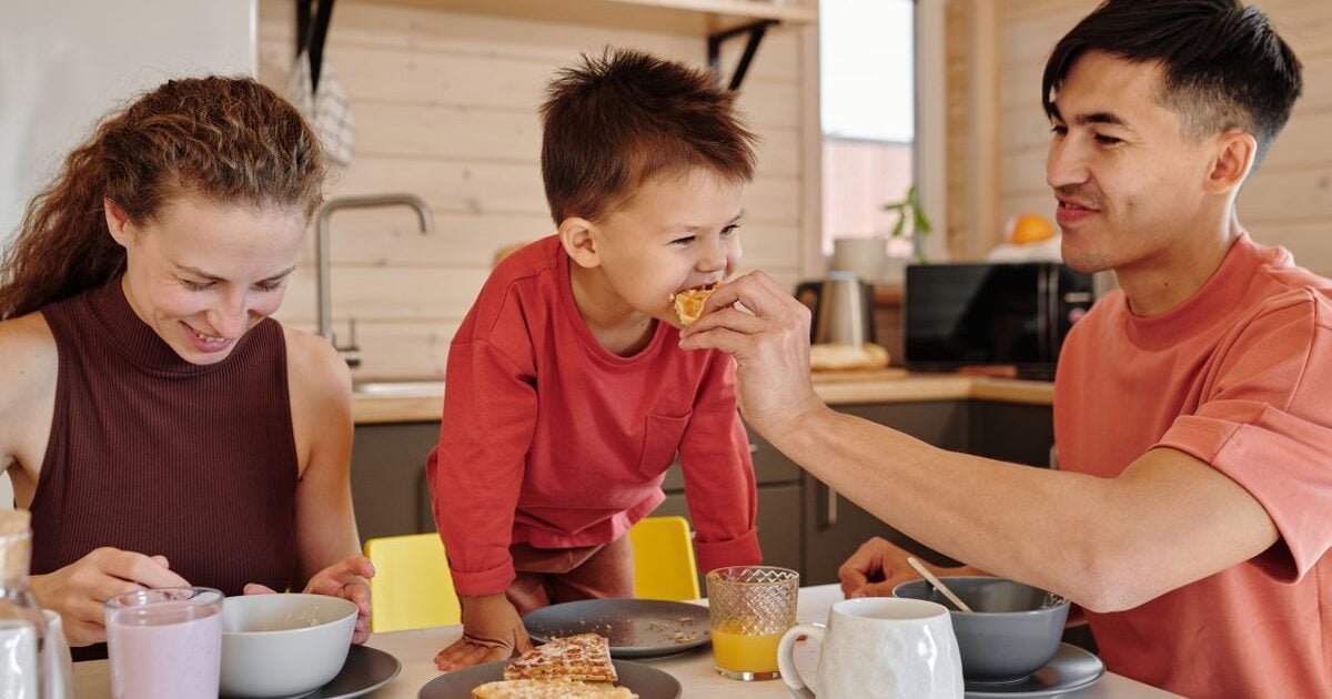 Parents and their young son eat breakfast at a kitchen table, with the father feeding the boy a waffle.