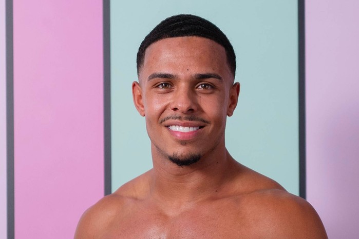 Ethan on Love Island season 13. A close up of a topless man smiling ahead in front of a light blue background.