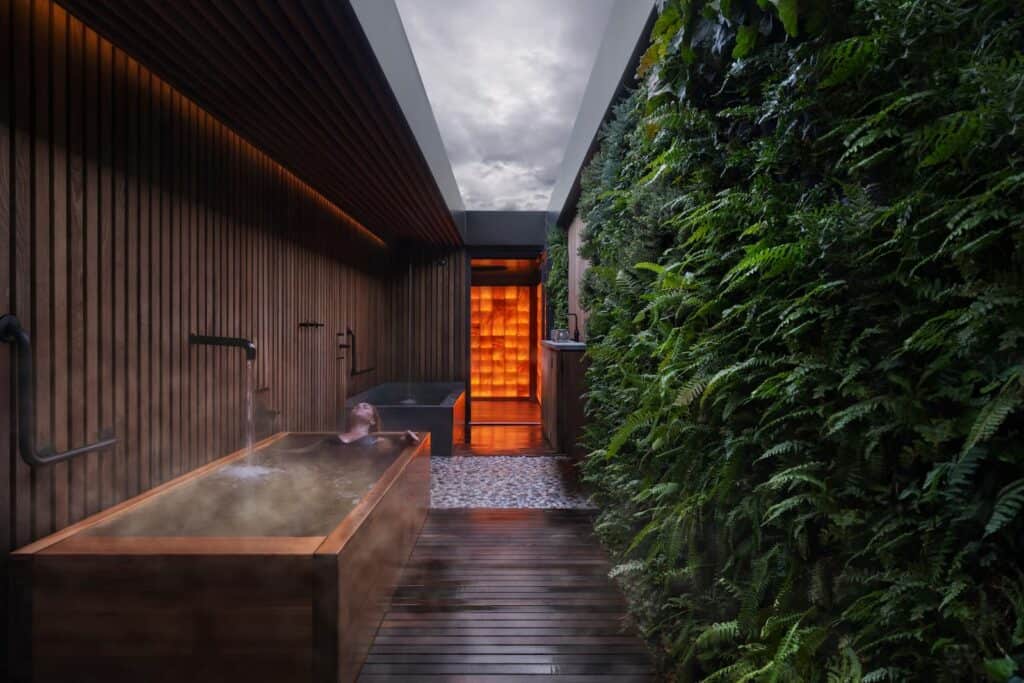 A modern indoor bath area with a wooden tub, lush green vertical garden wall, pebble floor, and a glowing orange-lit doorway in the background.