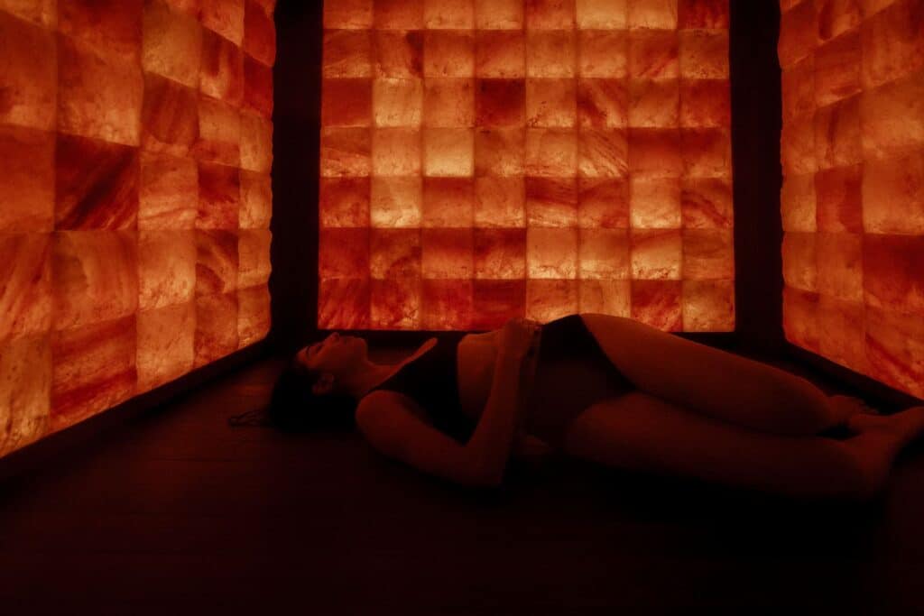 A person lies on the floor in a dim room with walls made of illuminated salt blocks, wearing dark clothing and facing upward.
