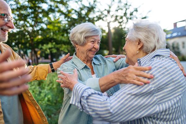 Two senior women greet each other warmly at an outdoor event.