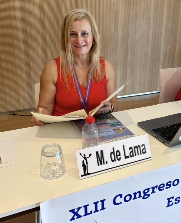 Alt text: A woman in a red sleeveless top sits behind a conference table, smiling while holding papers, with a nameplate reading “M. de Lama.”