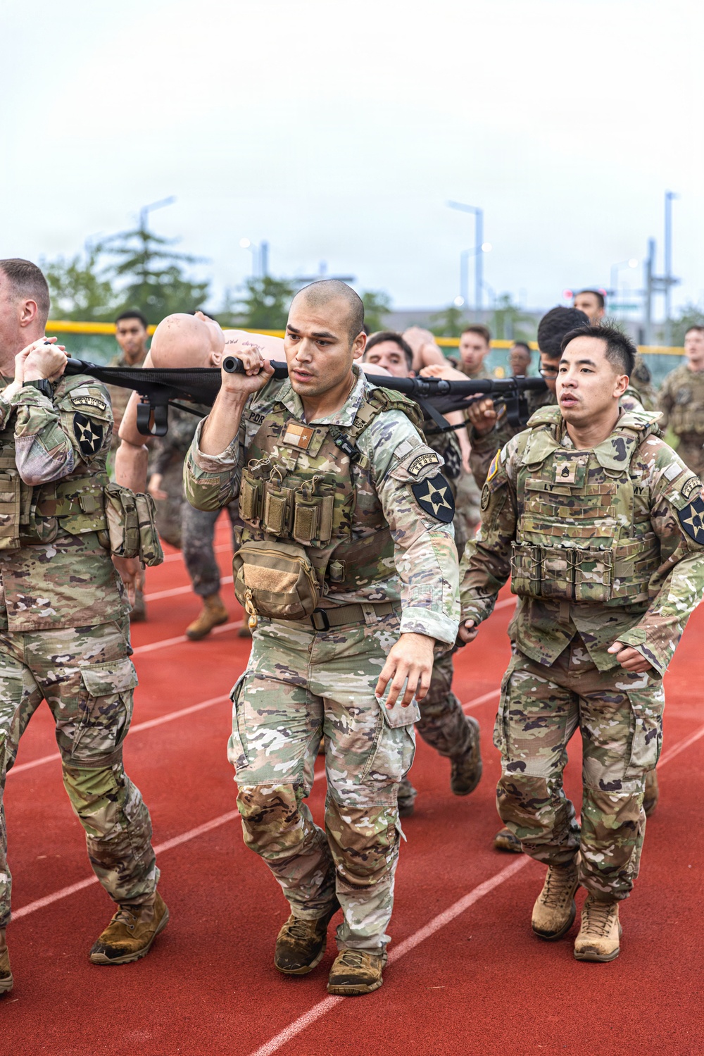 A group of U.S. Army soldiers in MultiCam (OCP) uniforms and plate carriers are conducting a litter carry drill on a red running track. They are transporting a training mannequin on a military stretcher/litter as part of what appears to be a combat casualty care or physical fitness exercise. Multiple soldiers wearing 2nd Infantry Division patches are visible, with additional personnel participating in the background.